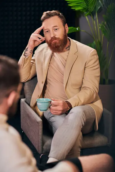 Gutaussehender bärtiger Mann mit Kaffeetasse sitzt während der Diskussion neben seinem Interviewer — Stockfoto