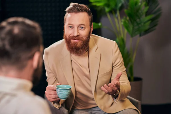 Hombres alegres en ropa de moda sentado con taza de café y discutir preguntas de la entrevista - foto de stock