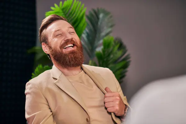 Guapo hombre alegre con barba en elegante atuendo riendo durante la entrevista mientras está en el estudio - foto de stock