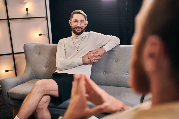 Hombres barbudos alegres en traje casual sonriendo y mirando a su entrevistador mientras está sentado en el estudio - foto de stock