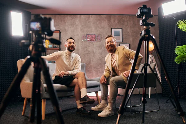 Hombres alegres de buen aspecto en trajes elegantes sonriendo felizmente a la cámara durante la entrevista en el estudio - foto de stock