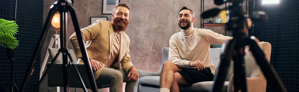 Красивые радостные мужчины в шикарных нарядах, счастливо улыбающиеся в камеру во время интервью в студии, баннер — стоковое фото