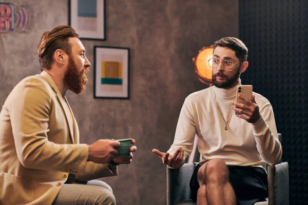 Due bei uomini eleganti con la barba in abiti chic guardando smartphone durante l'intervista — Foto stock