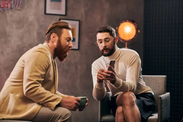 Deux beaux hommes élégants avec barbe dans des vêtements chics en regardant smartphone pendant l'interview — Photo de stock