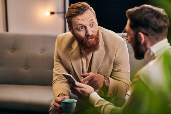 Hombre barbudo guapo hablando con su entrevistador en gafas que sostiene el teléfono inteligente durante la discusión - foto de stock
