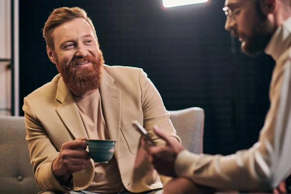 Lustiger rothaariger Mann im Gespräch mit seinem Interviewer mit einer Brille, die während der Diskussion das Smartphone hält — Stockfoto