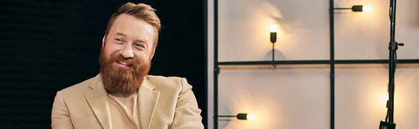 Alegre hombre guapo con barba roja en ropa elegante sentado y sonriendo durante la entrevista, pancarta - foto de stock