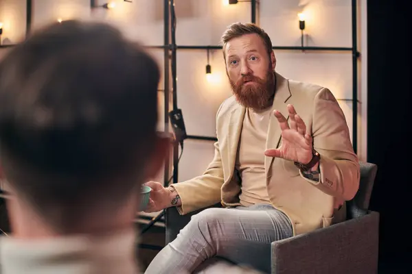 Bel homme roux avec barbe dans des vêtements élégants parler avec son intervieweur tout en studio — Photo de stock