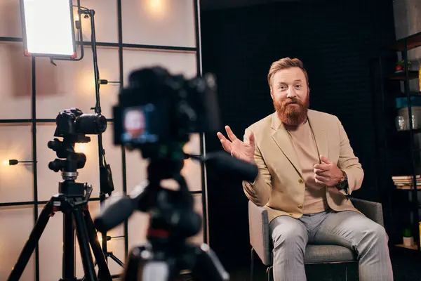 Веселый человек с рыжими волосами и бородой в элегантном наряде активно говорит во время подкаста в студии — стоковое фото