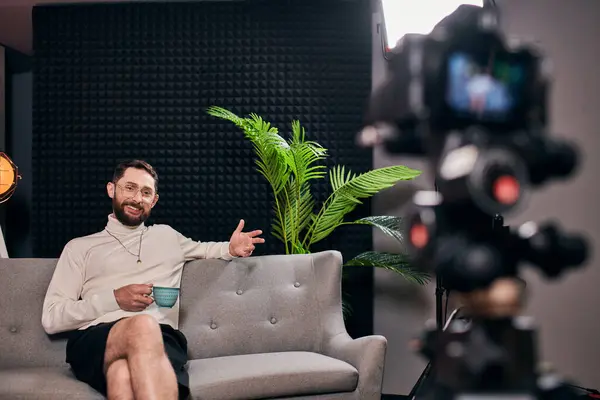 Allegro uomo barbuto in elegante abbigliamento con bicchieri in possesso di tazza di caffè durante l'intervista in studio — Foto stock