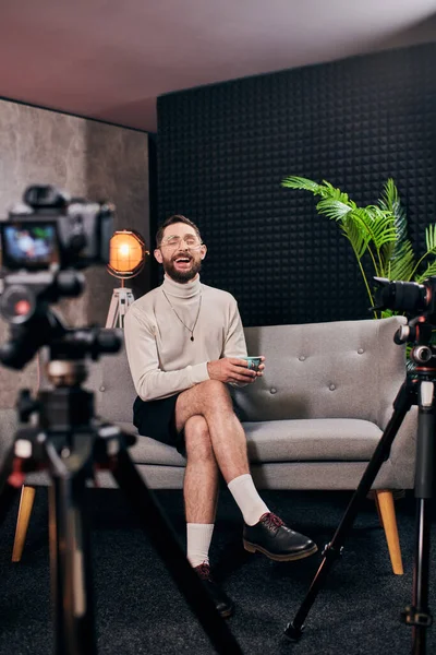 Cheerful bearded man in elegant attire with glasses holding coffee cup during interview in studio — Stock Photo