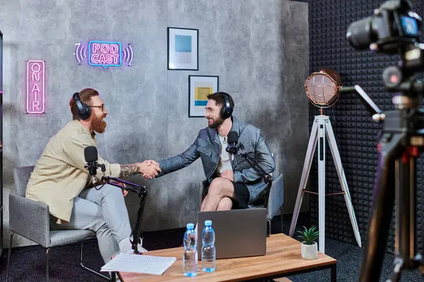 Homens barbudos de boa aparência em trajes casuais confortáveis com fones de ouvido apertando as mãos durante o podcast — Fotografia de Stock