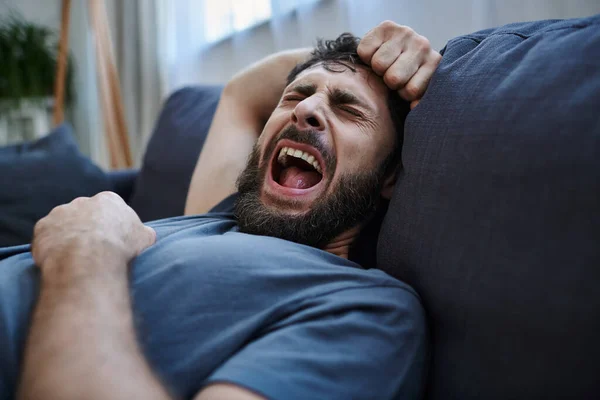 Desesperado hombre deprimido en ropa de casa en el sofá gritando durante el colapso, conciencia de salud mental - foto de stock