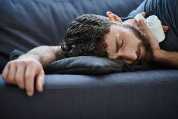 Homem sofredor traumatizado com barba deitada no sofá com pílulas na mão, consciência de saúde mental — Fotografia de Stock