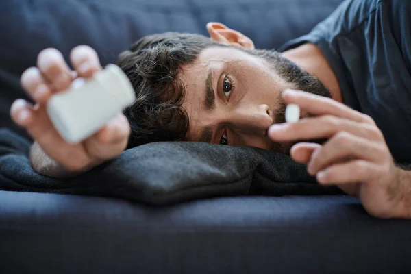 Ill suffering man in casual attire lying on sofa with pills in hand, mental health awareness — Stock Photo