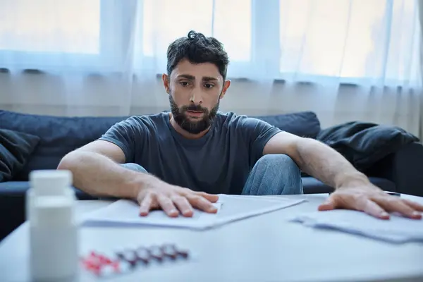 Depressiver kranker Mann mit Bart sitzt während depressiver Episode mit Papieren und Tabletten am Tisch — Stockfoto