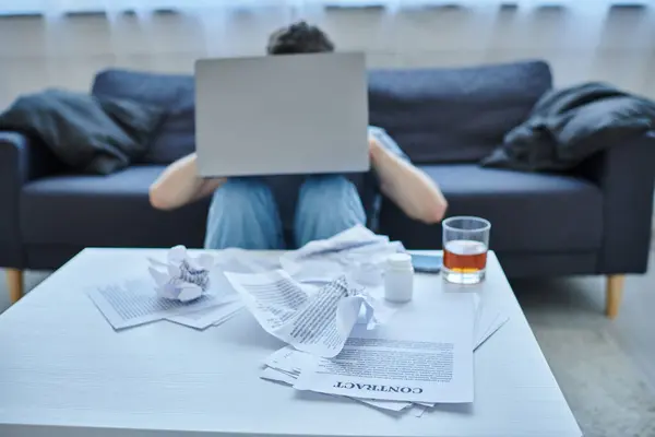 Hombre traumatizado deprimido con barba trabajando en el ordenador portátil con un vaso de bebida alcohólica en la mesa - foto de stock