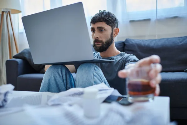 Homme traumatisé déprimé avec barbe travaillant à l'ordinateur portable avec un verre de boisson alcoolisée sur la table — Photo de stock