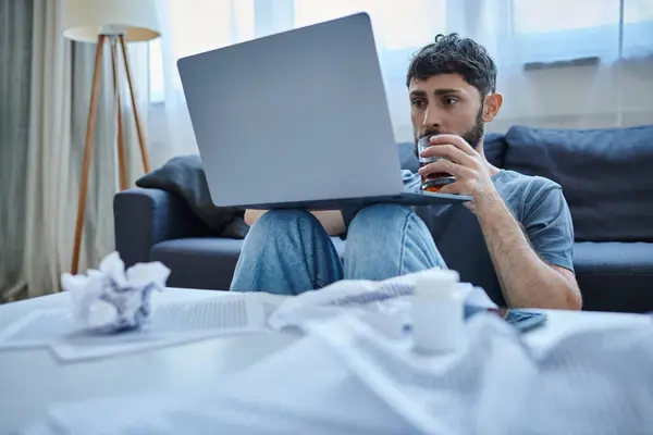Desperate traumatized man with beard working at laptop with and drinking alcohol drink on table — Stock Photo