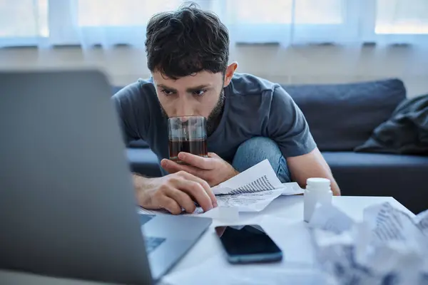 Verzweifelter traumatisierter Mann mit Bart arbeitet am Laptop und trinkt Alkohol auf dem Tisch — Stockfoto