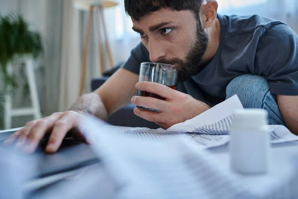Hombre traumatizado desesperado con barba trabajando en el ordenador portátil con y beber alcohol en la mesa - foto de stock