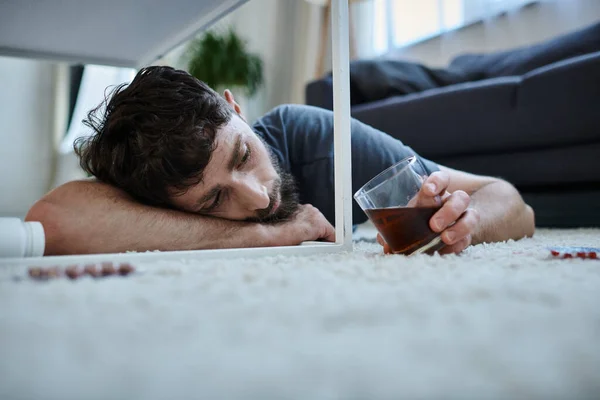 Депресивний чоловік в повсякденному вбранні п'є алкоголь і дивиться на таблетки під час психічного розладу — стокове фото