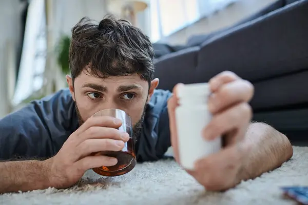 Расстроенный больной человек в повседневной одежде пьет алкоголь и держит таблетки во время психического срыва — стоковое фото