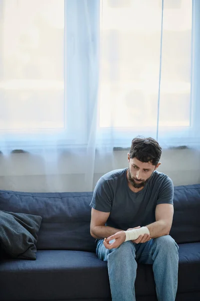 Depressed man with bandage on arm after attempting suicide sitting on sofa, mental health awareness — Stock Photo