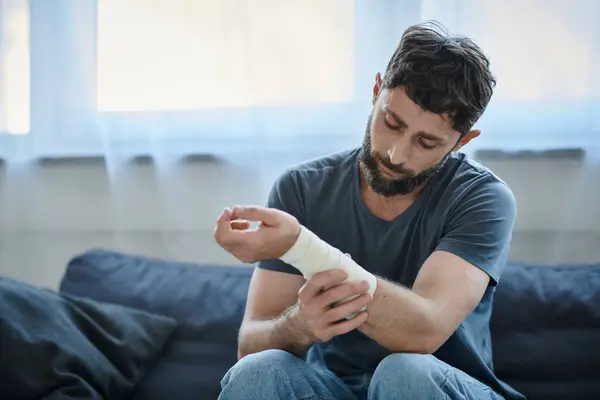 Hombre deprimido con vendaje en el brazo después de intentar suicidarse sentado en el sofá, conciencia de salud mental - foto de stock