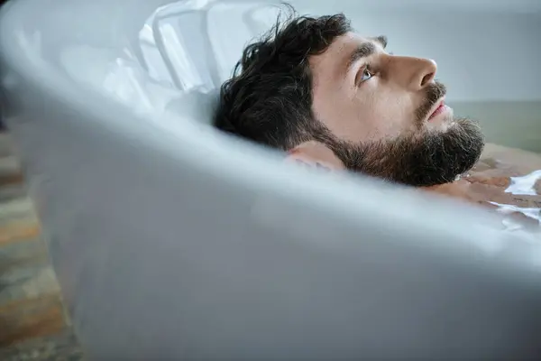 Hombre frustrado deprimido con barba acostado en la bañera durante la ruptura, conciencia de salud mental - foto de stock