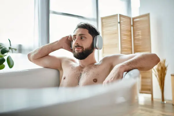Allegro bell'uomo con barba e cuffie seduto e rilassante nella sua vasca da bagno, salute mentale — Foto stock