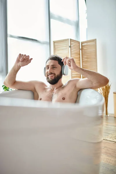 Cheerful handsome man with beard and headphones sitting and relaxing in his bathtub, mental health — Stock Photo