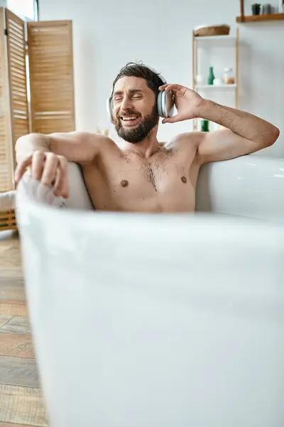 Веселый привлекательный мужчина с бородой и наушниками сидит и расслабляется в ванной, психическое здоровье — стоковое фото