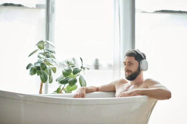Jolly good looking man with beard and headphones sitting and relaxing in his bathtub, mental health — Stock Photo