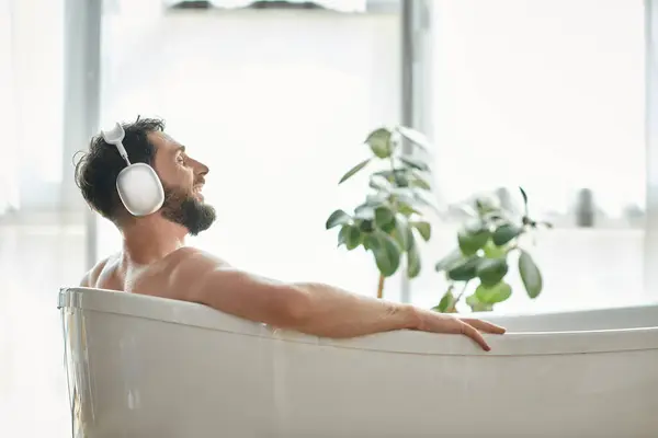 Fröhlich gut aussehender Mann mit Bart und Kopfhörer sitzt und entspannt in seiner Badewanne, psychische Gesundheit — Stockfoto