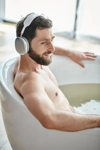 Jolly good looking man with beard and headphones sitting and relaxing in his bathtub, mental health — Stock Photo