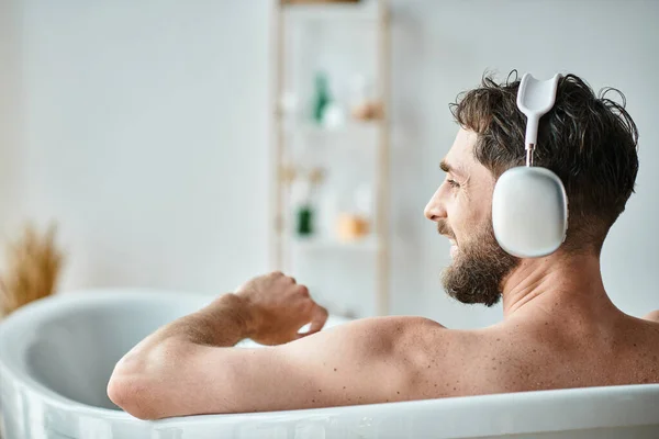Merry handsome man with beard and headphones sitting and relaxing in his bathtub, mental health — Stock Photo