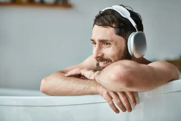Homem bonito alegre com barba e fones de ouvido sentado e relaxante em sua banheira, saúde mental — Fotografia de Stock