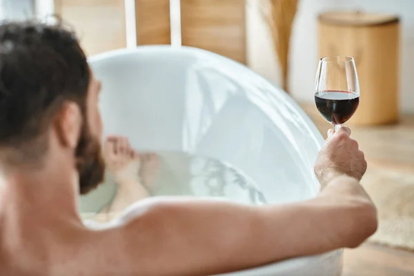 Сосредоточьтесь на бокале красного вина в руках размытого расслабляющего бородатого человека в ванной, психического здоровья — стоковое фото