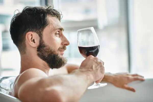 Gioioso bell'uomo con barba rilassante nella vasca da bagno con bicchiere di vino rosso, consapevolezza della salute mentale — Foto stock