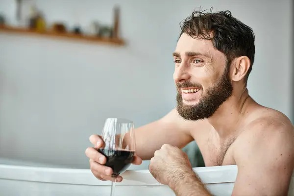 Happy attractive man with beard relaxing in bathtub with glass of red wine, mental health awareness — Stock Photo