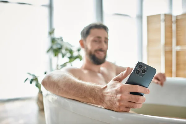 Concentrarsi sul moderno smartphone nelle mani di uomo barbuto allegro offuscata fare il bagno, la salute mentale — Foto stock