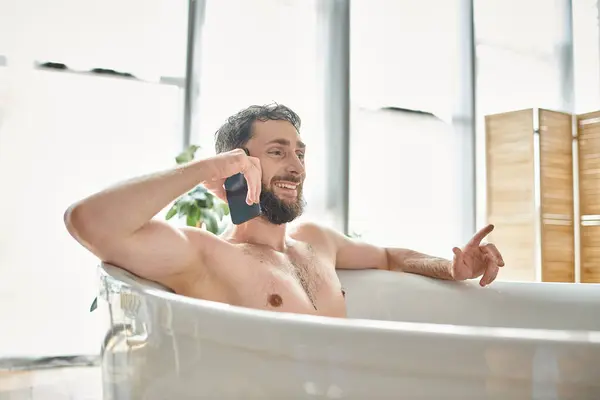 Freudig gut aussehender Mann mit Bart telefoniert, während er sich in der Badewanne entspannt, psychische Gesundheit — Stockfoto