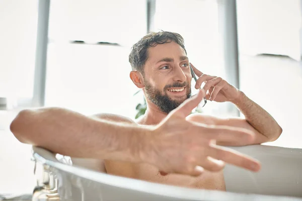 Gioioso bell'uomo con la barba che parla per telefono mentre si rilassa nella vasca da bagno, salute mentale — Foto stock