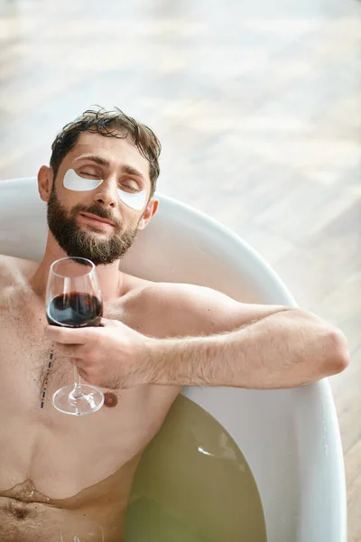 Веселый привлекательный мужчина с бородой и повязками на глазу расслабляется в ванне с бокалом красного вина — стоковое фото