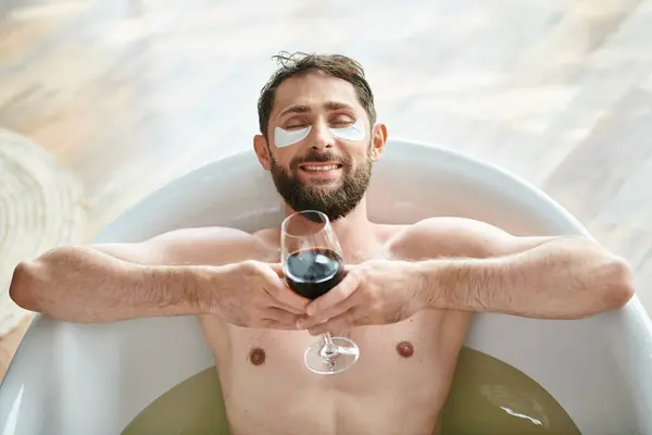 Joyeux homme attrayant avec la barbe et les yeux patchs relaxant dans la baignoire avec un verre de vin rouge — Photo de stock