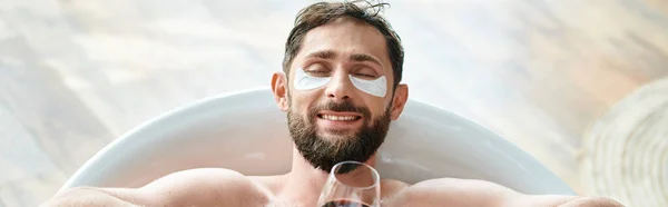 Весело привлекательный мужчина с бородой и повязками на глазу расслабляется в ванне с бокалом красного вина, баннер — стоковое фото
