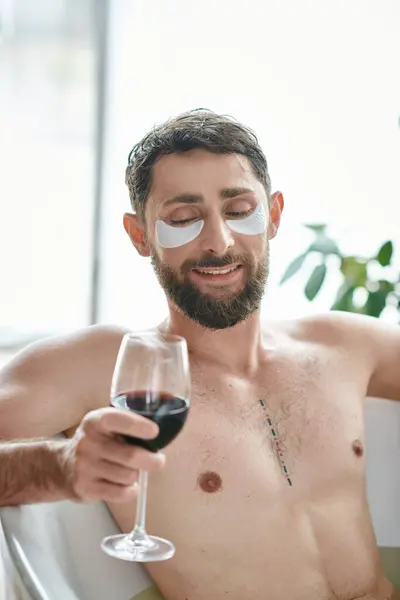Fröhlicher gutaussehender Mann mit Bart und Augenklappen entspannt in der Badewanne bei einem Glas Rotwein — Stockfoto