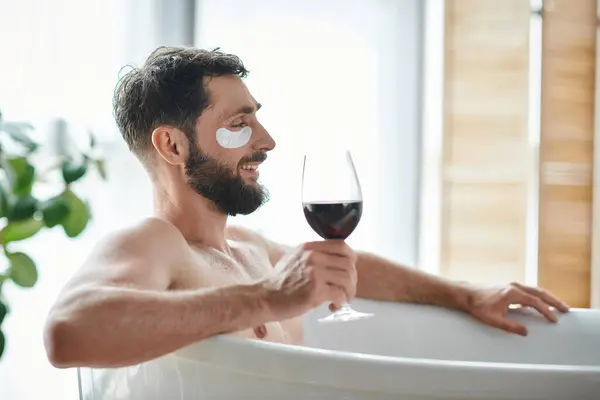 Весёлый, красивый мужчина с бородой и повязками на глазу расслабляется в ванной с бокалом красного вина — стоковое фото