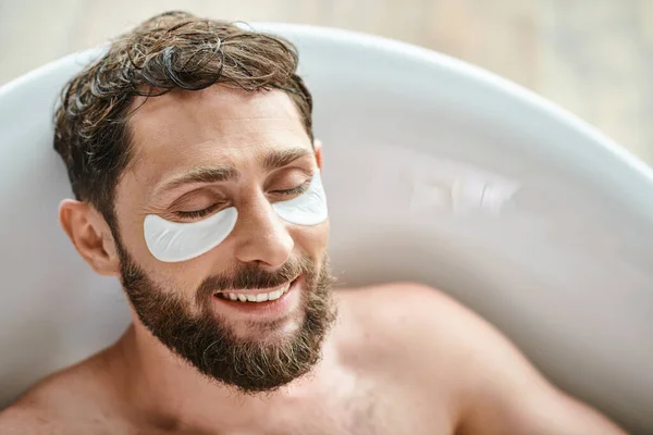 Allegro bell'uomo con barba rilassante nella sua vasca da bagno con macchie sugli occhi, salute mentale — Foto stock